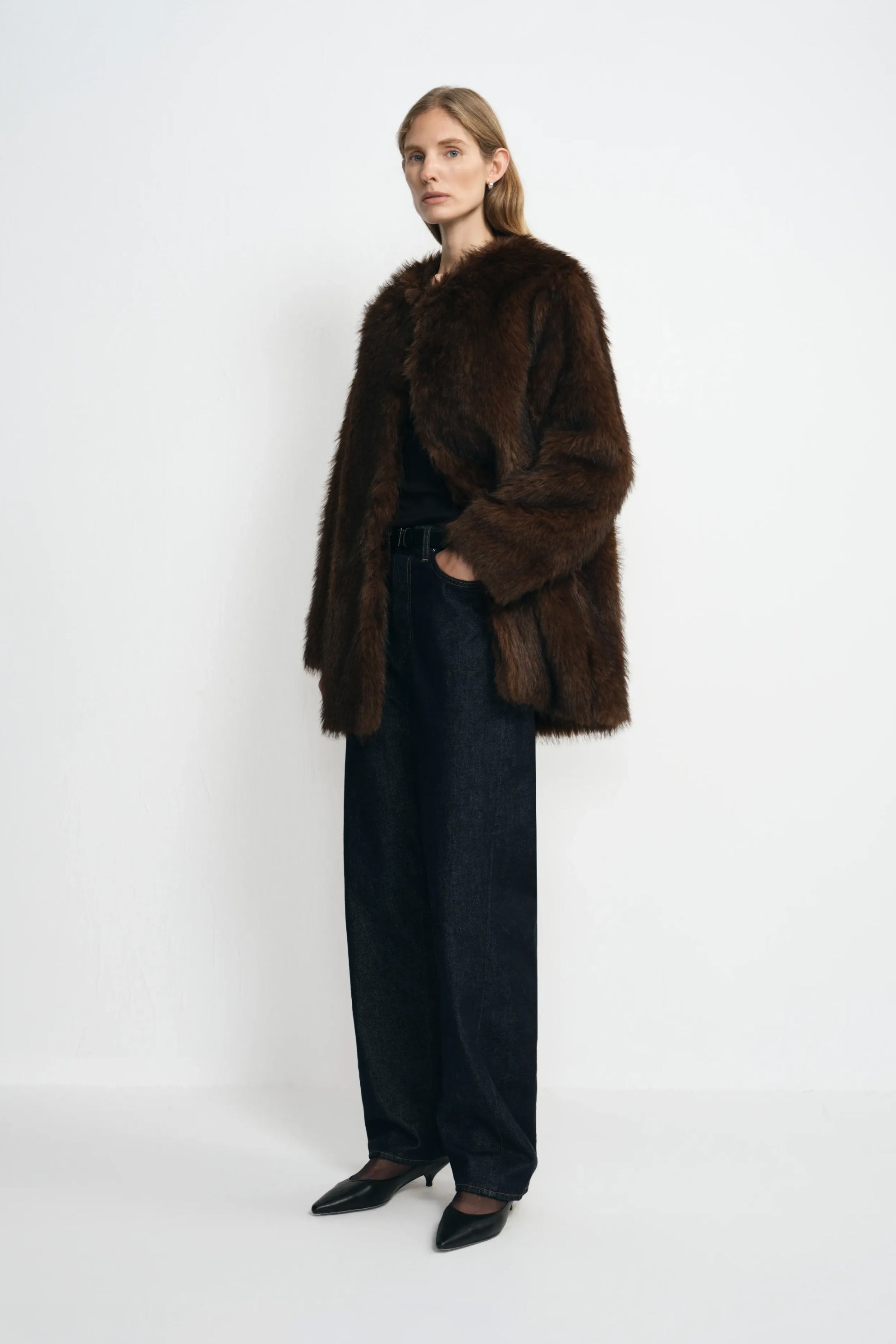 A model standing at an angle in a brown collarless faux fur coat with full sleeves, worn open over a black top and dark jeans with black pointed shoes.