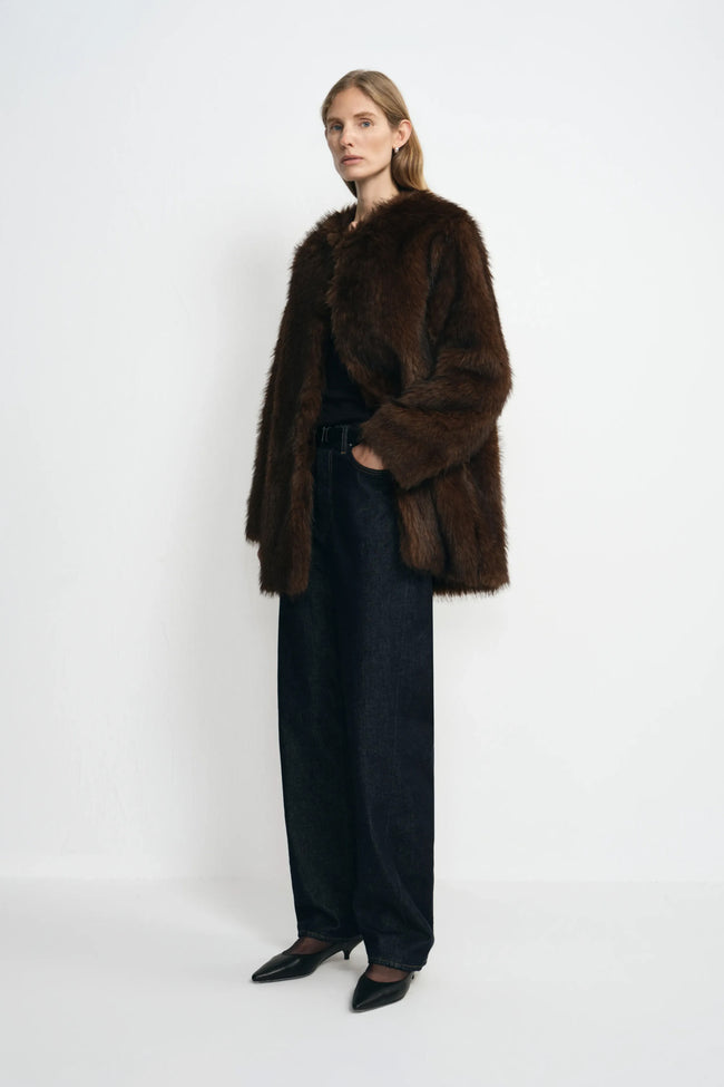 A model standing at an angle in a brown collarless faux fur coat with full sleeves, worn open over a black top and dark jeans with black pointed shoes.