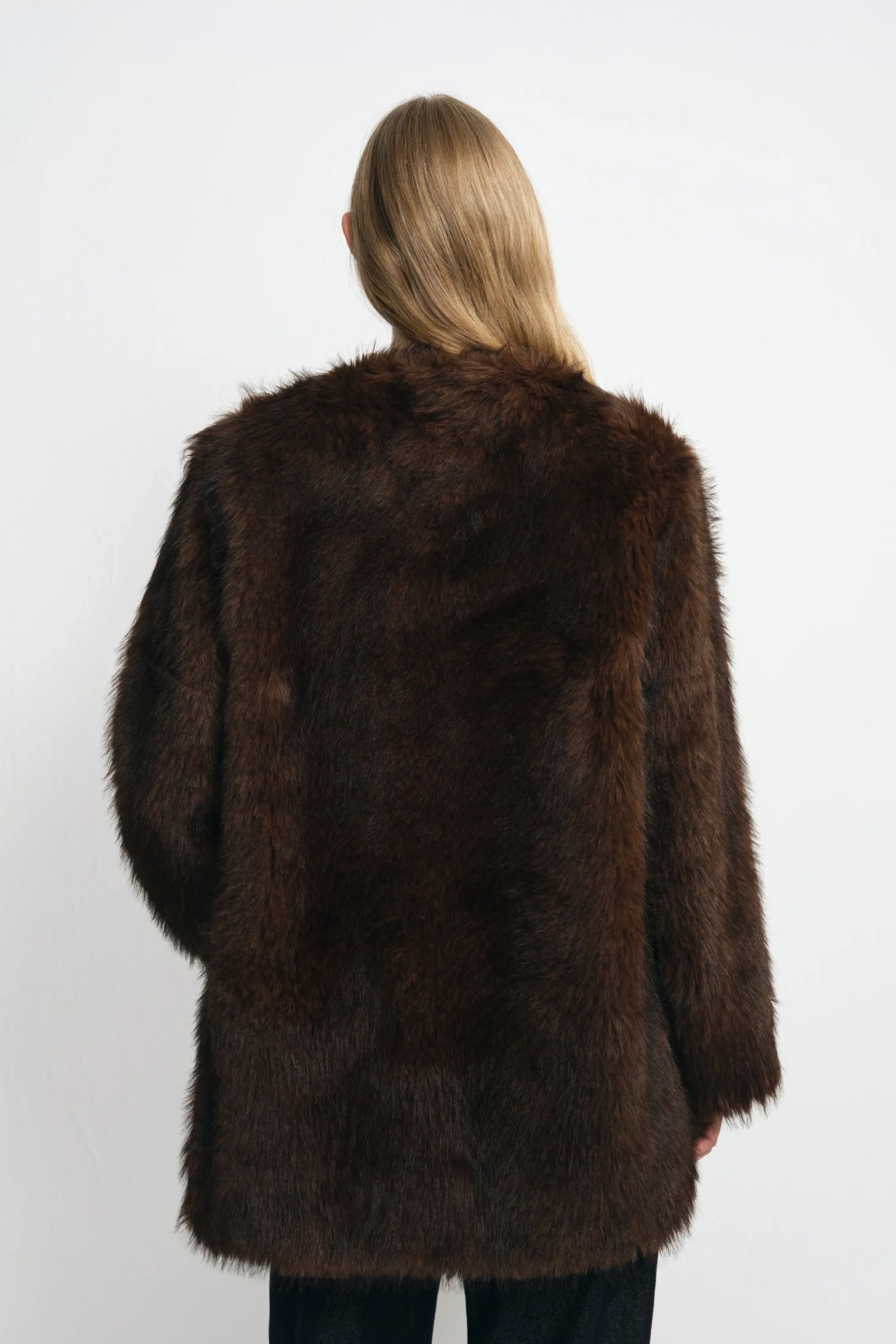 Back view of a brown faux fur coat on a model, showing the long-pile texture across the shoulders, back and broad sleeves above dark jeans.