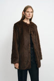 Three-quarter close-up of a model in a brown faux fur coat, showing the collarless neckline and full sleeves over a black top and belted dark jeans.