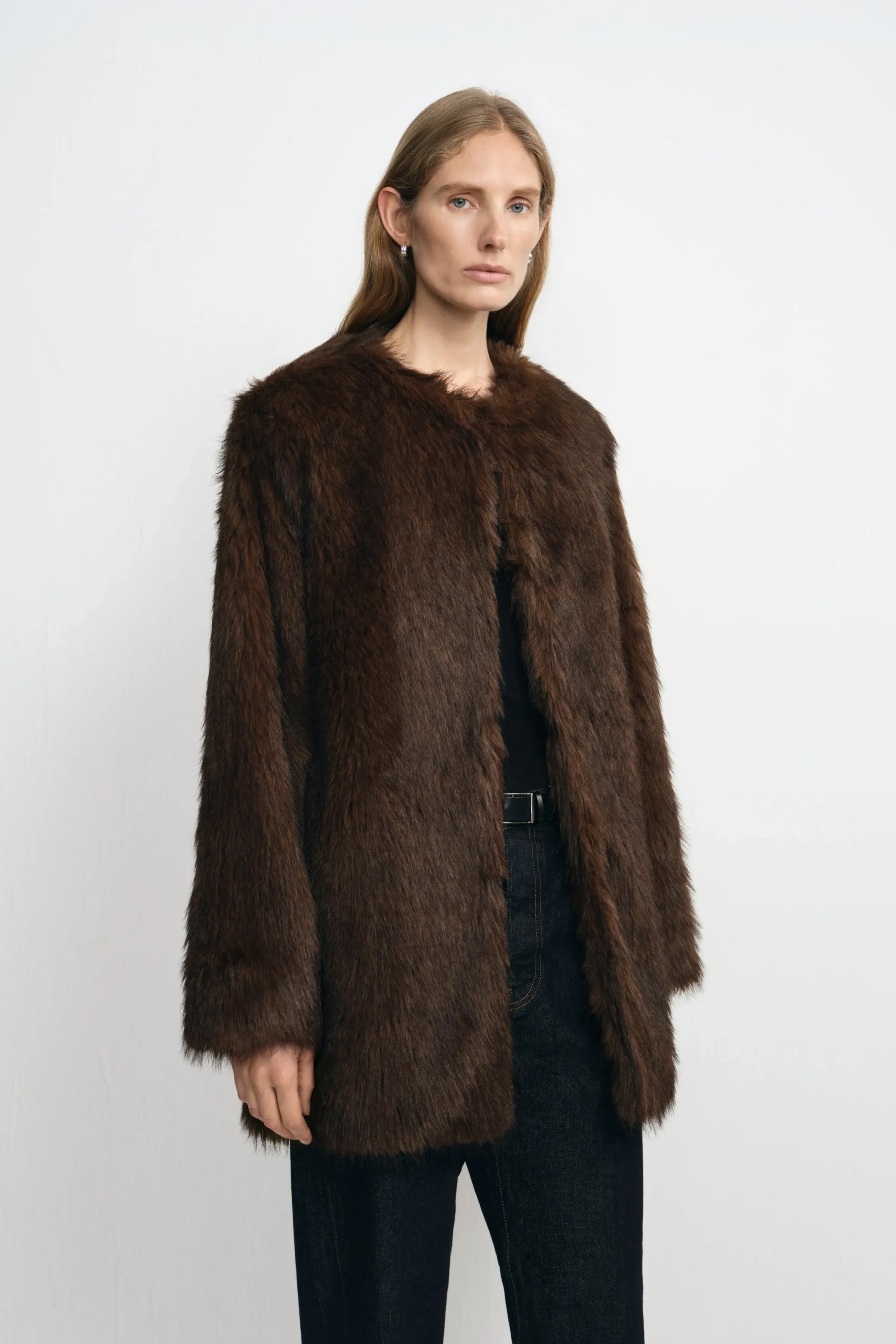 Three-quarter close-up of a model in a brown faux fur coat, showing the collarless neckline and full sleeves over a black top and belted dark jeans.