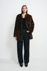 A model wearing a brown faux fur coat with a round collarless neckline, open over a black tank and dark jeans, paired with pointed black shoes.