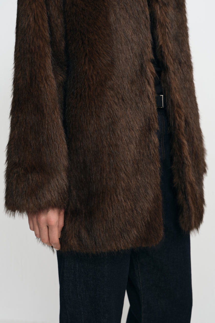 Close-up of a brown faux fur coat on a model, showing the long-pile texture, wide sleeve and straight front edge over dark jeans.