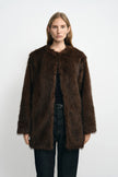Front view of a model wearing a brown faux fur coat with a collarless round neck and straight front edges, layered over a black top and belted jeans.