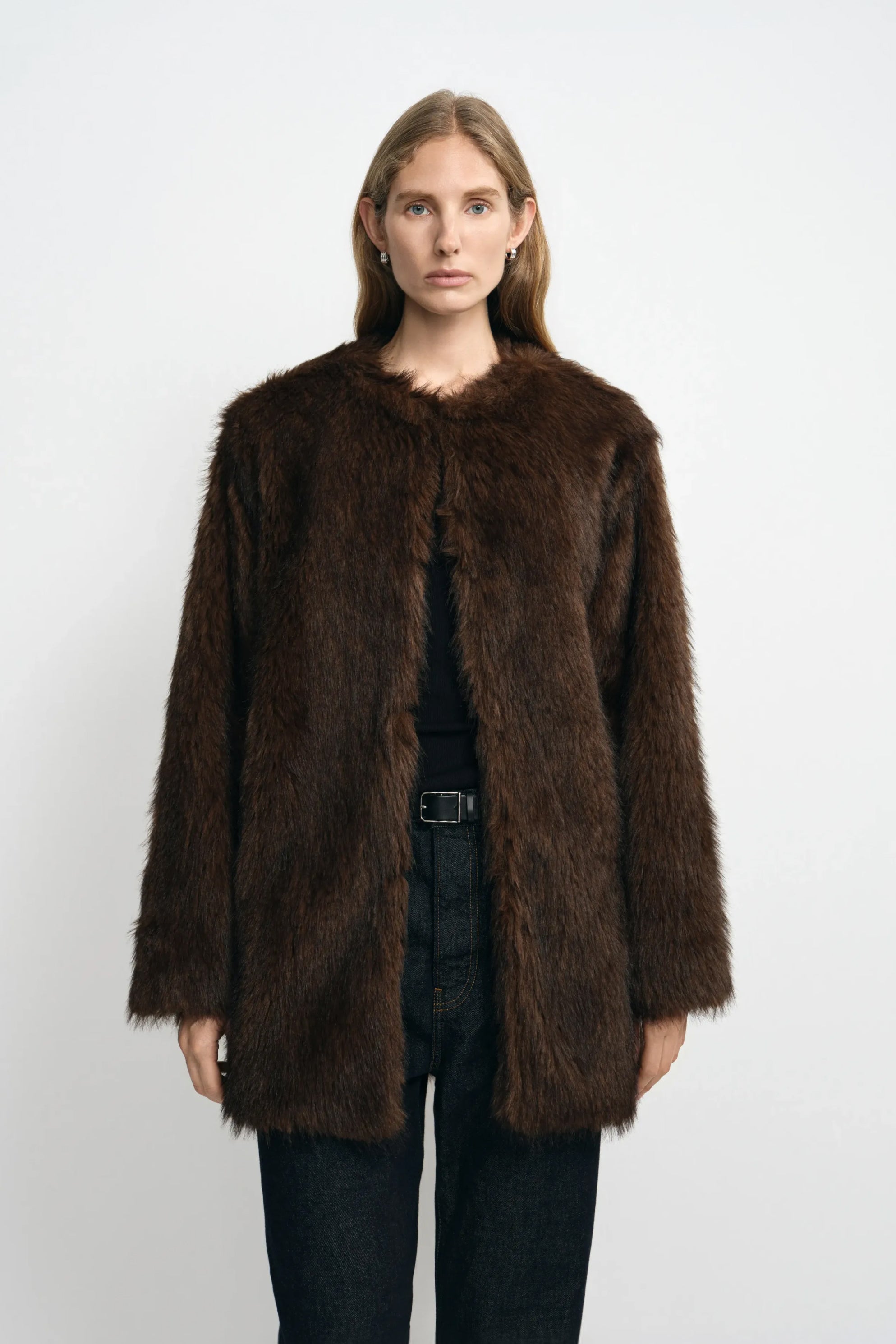 Front view of a model wearing a brown faux fur coat with a collarless round neck and straight front edges, layered over a black top and belted jeans.