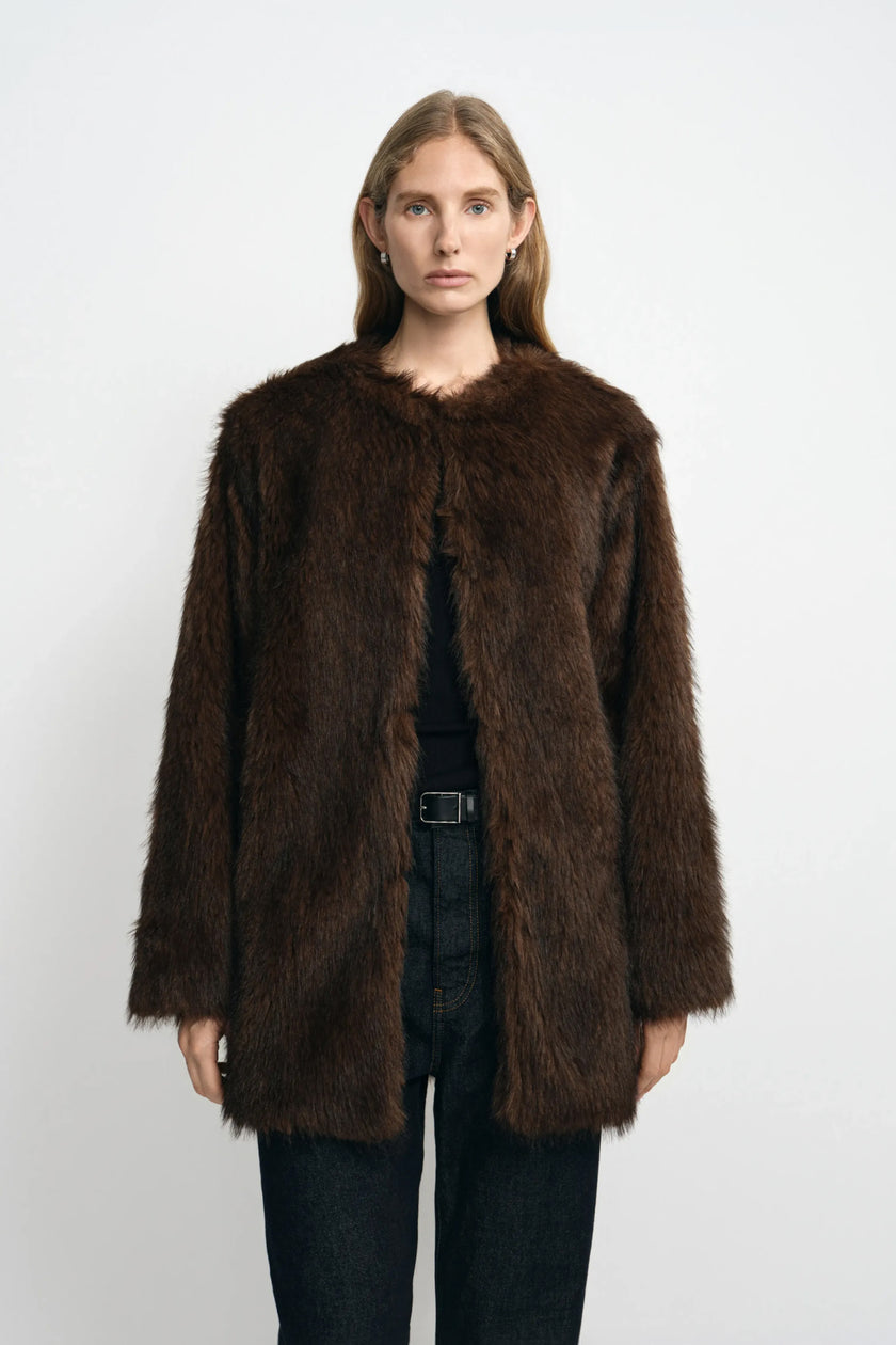 Front view of a model wearing a brown faux fur coat with a collarless round neck and straight front edges, layered over a black top and belted jeans.