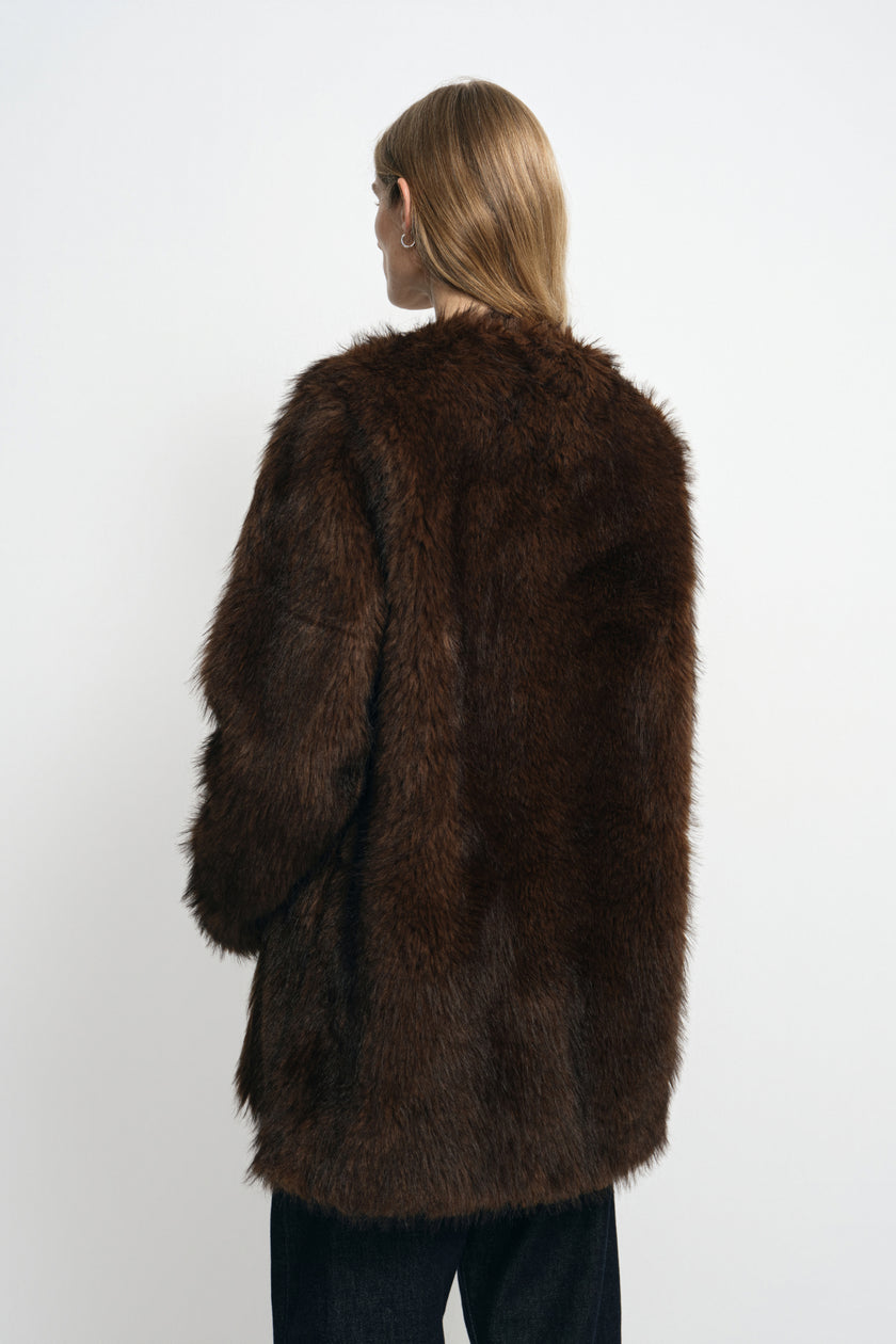 Rear three-quarter view of a model wearing a brown faux fur coat, showing its long-pile surface, rounded shoulders and straight hem above dark jeans.