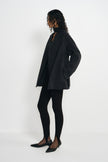Side view of a model in an open heather-grey short coat with long sleeves and a straight hem, worn with a dark scarf, black leggings and pointed shoes.
