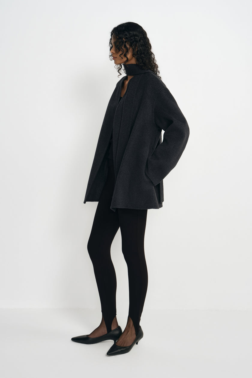 Side view of a model in an open heather-grey short coat with long sleeves and a straight hem, worn with a dark scarf, black leggings and pointed shoes.
