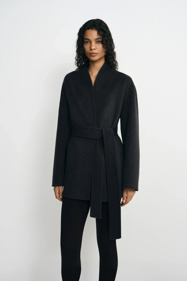 Front detail of a model wearing a heather-grey wrap coat, with the self-fabric belt tied at the waist and long ends falling over black leggings.