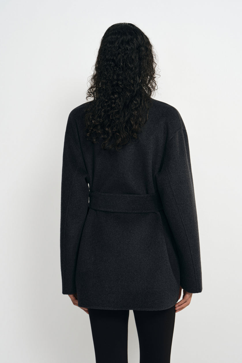 Back view of a heather-grey short coat on a model, showing the belt across the waist, long sleeves and straight hem above black leggings.