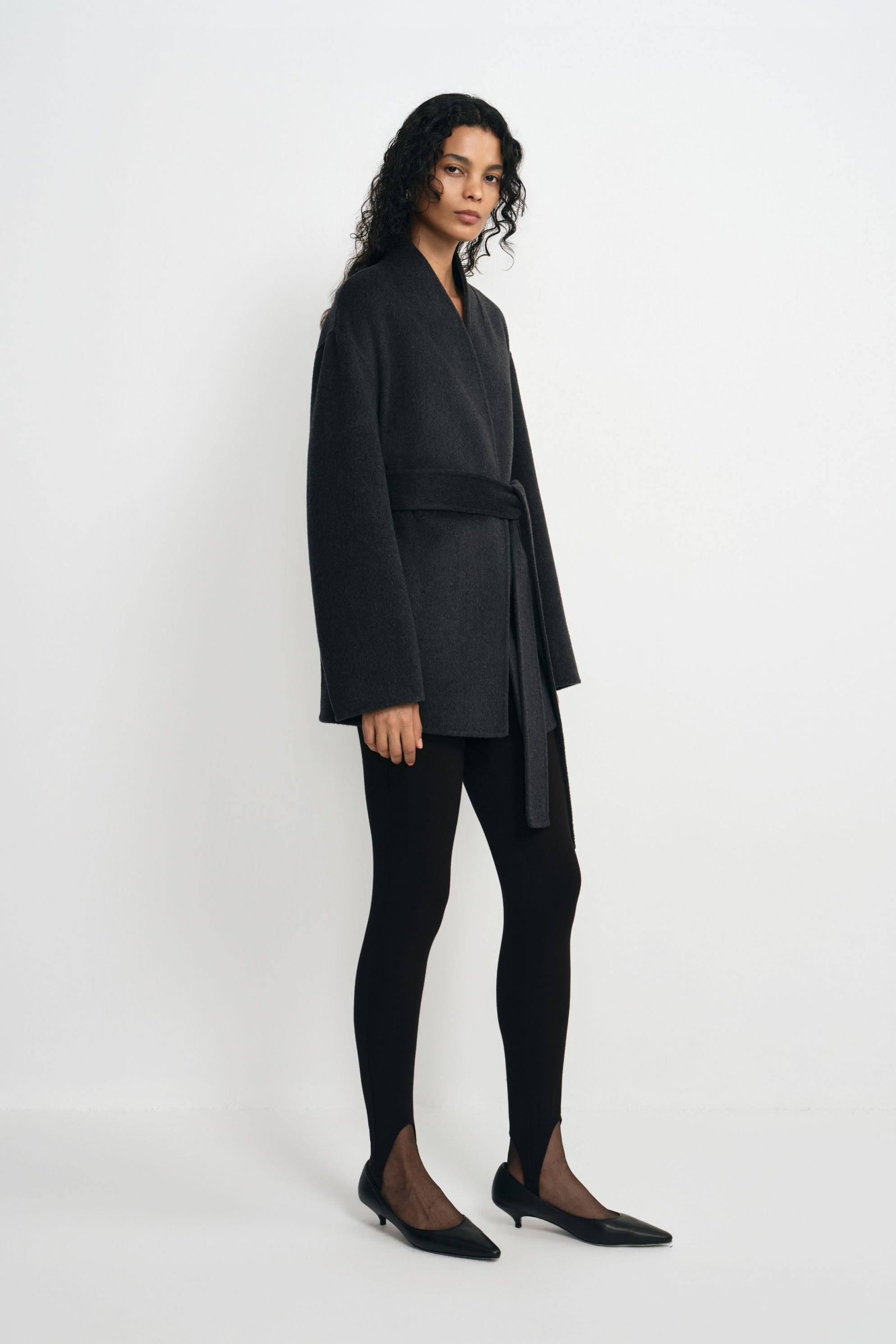 Three-quarter view of a model in a heather-grey belted wrap coat with a V-shaped opening, paired with black leggings and pointed black shoes.