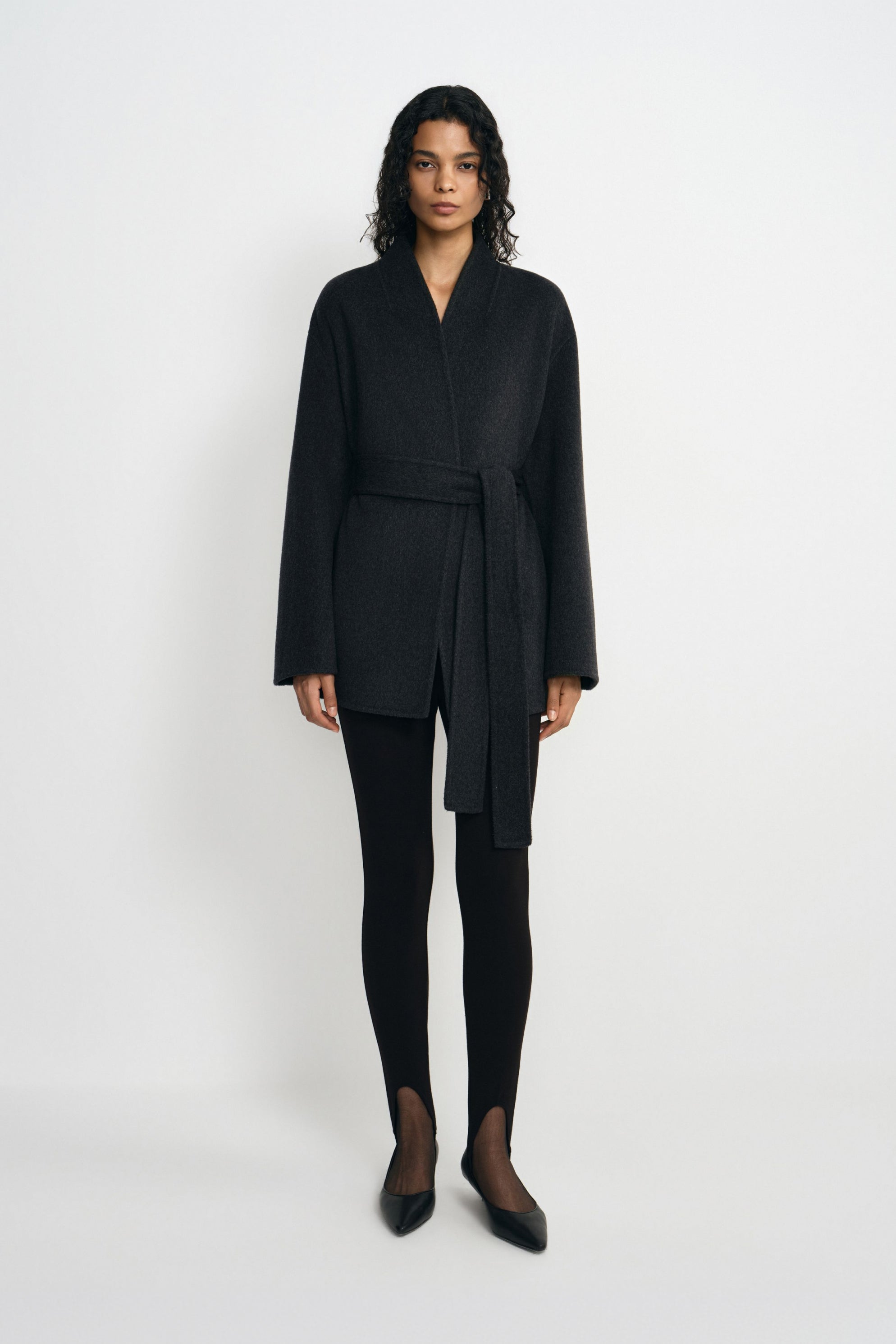 A model facing forward in a heather-grey wool-alpaca-blend short coat tied at the waist, styled with black split-hem leggings and pointed shoes.