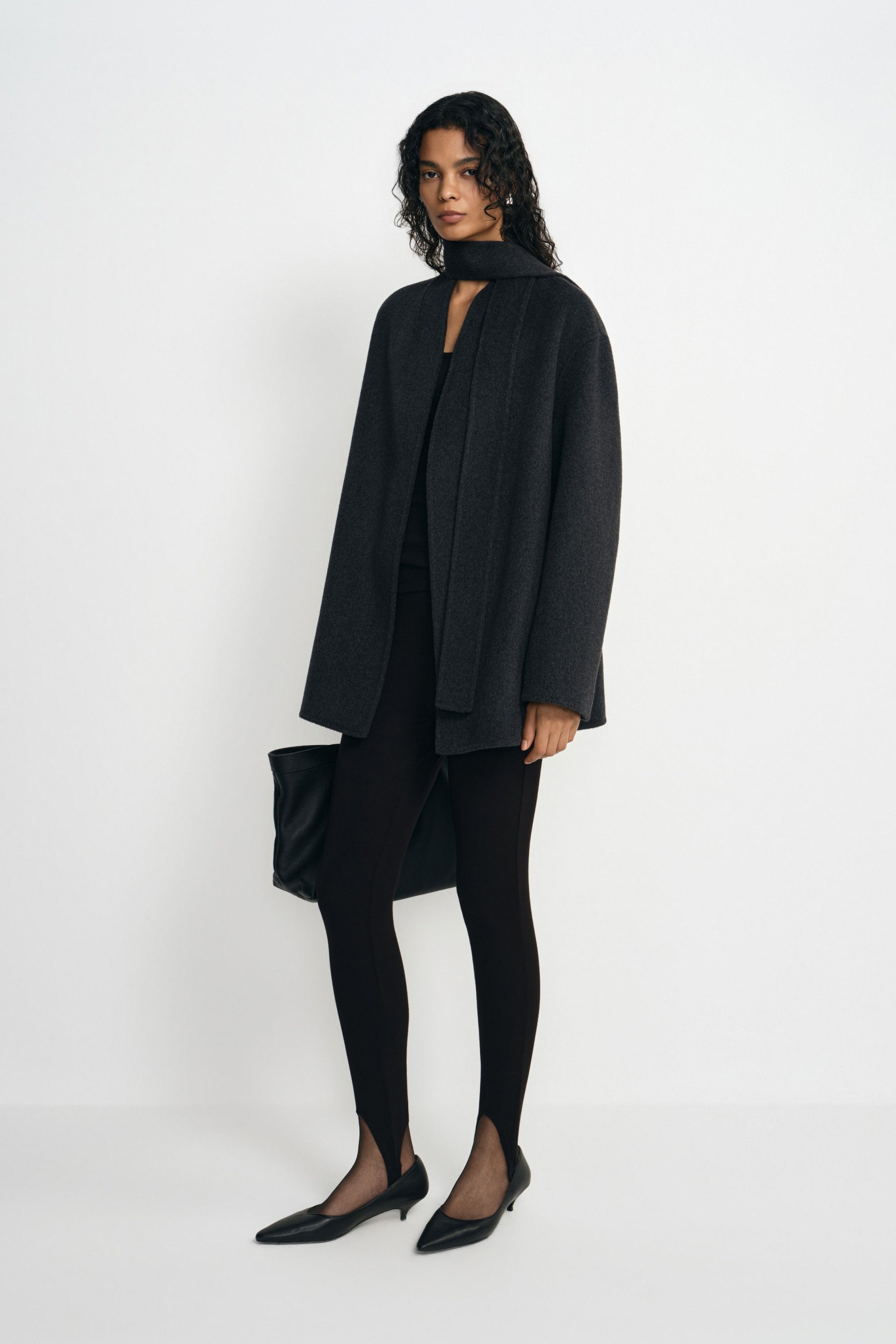 A model wearing a heather-grey wool-alpaca-blend short coat open with a dark neck scarf, paired with black leggings, pointed shoes and a black bag.