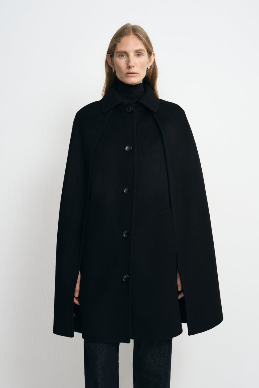 Front view of a model in a black cape coat, showing the pointed collar, row of buttons and wide drape around the arms above dark trousers.