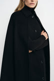 Close-up of a black cape coat on a model, showing the pointed collar and front buttons, with a black turtleneck sleeve visible through an arm opening.