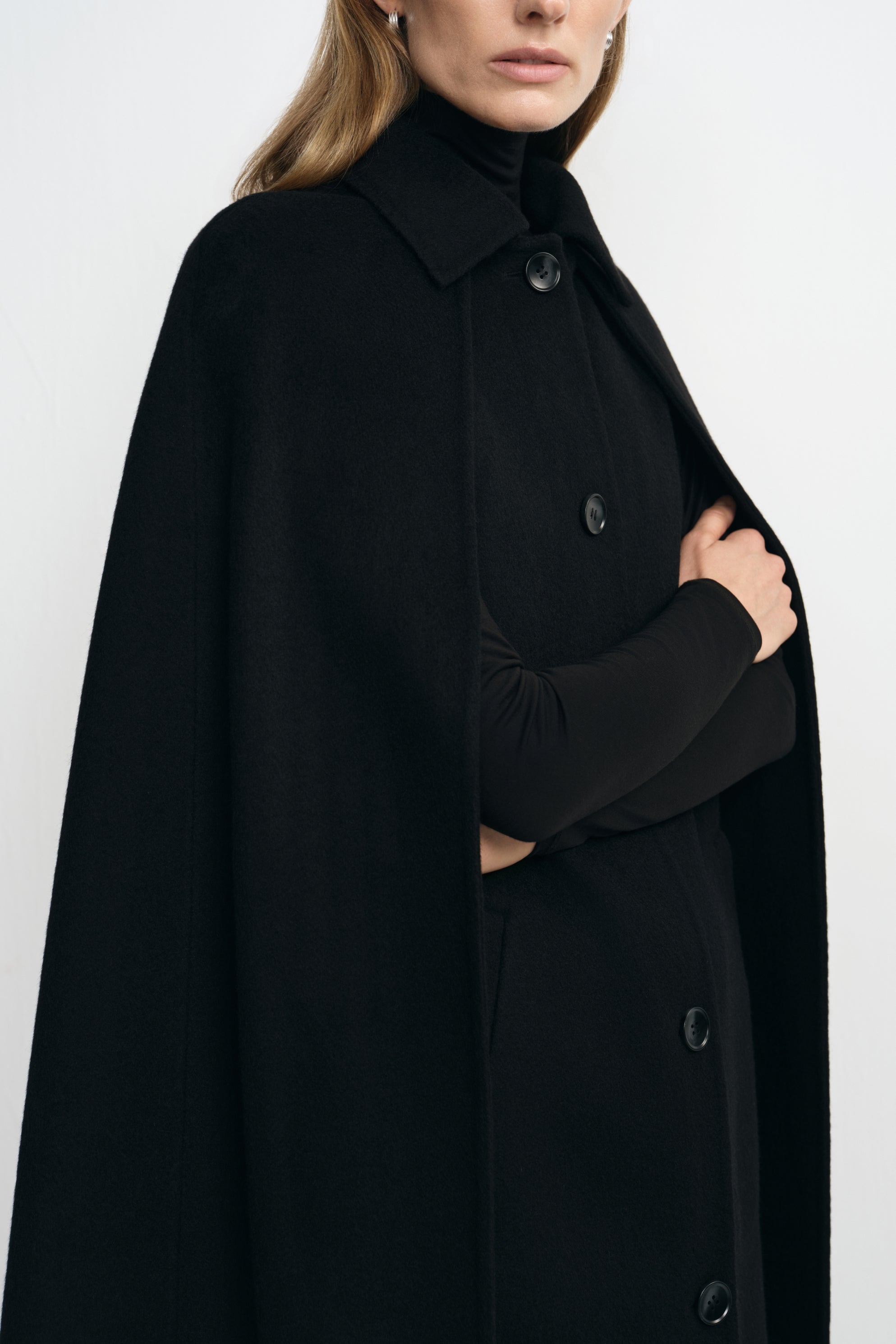 Close-up of a black cape coat on a model, showing the pointed collar and front buttons, with a black turtleneck sleeve visible through an arm opening.