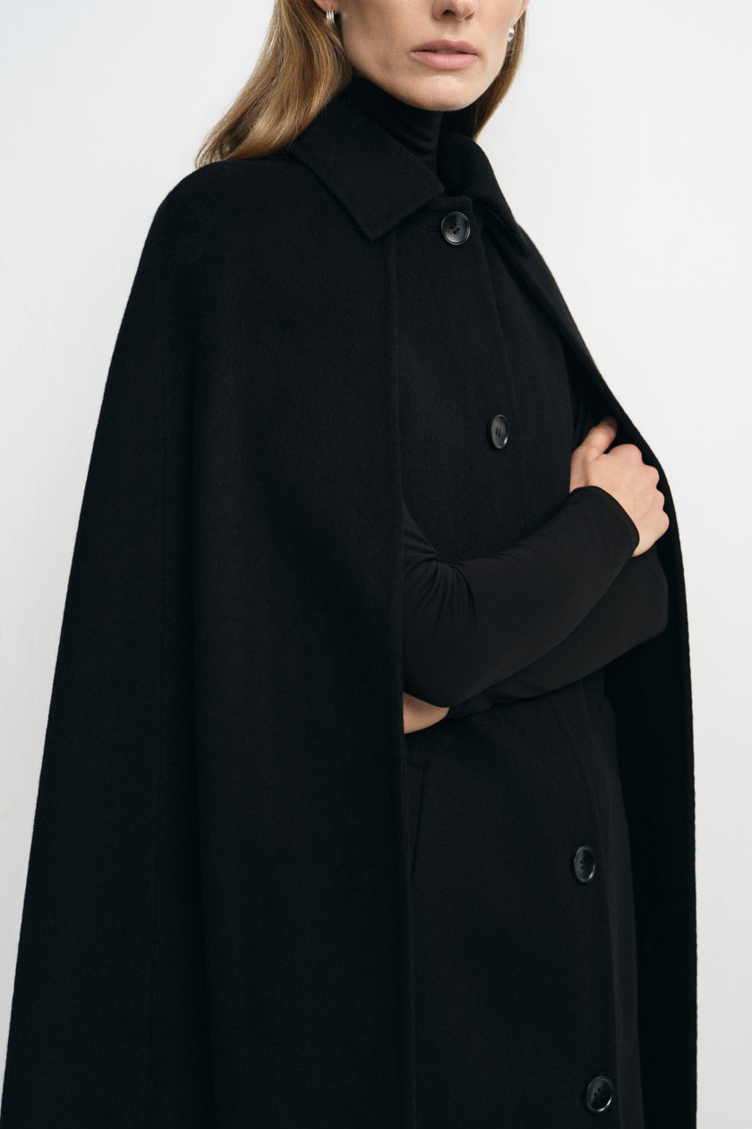 Close-up of a black cape coat on a model, showing the pointed collar and front buttons, with a black turtleneck sleeve visible through an arm opening.