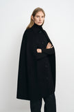 Three-quarter view of a model in a black cape coat, with folded arms emerging from the openings beneath the pointed collar and button front.