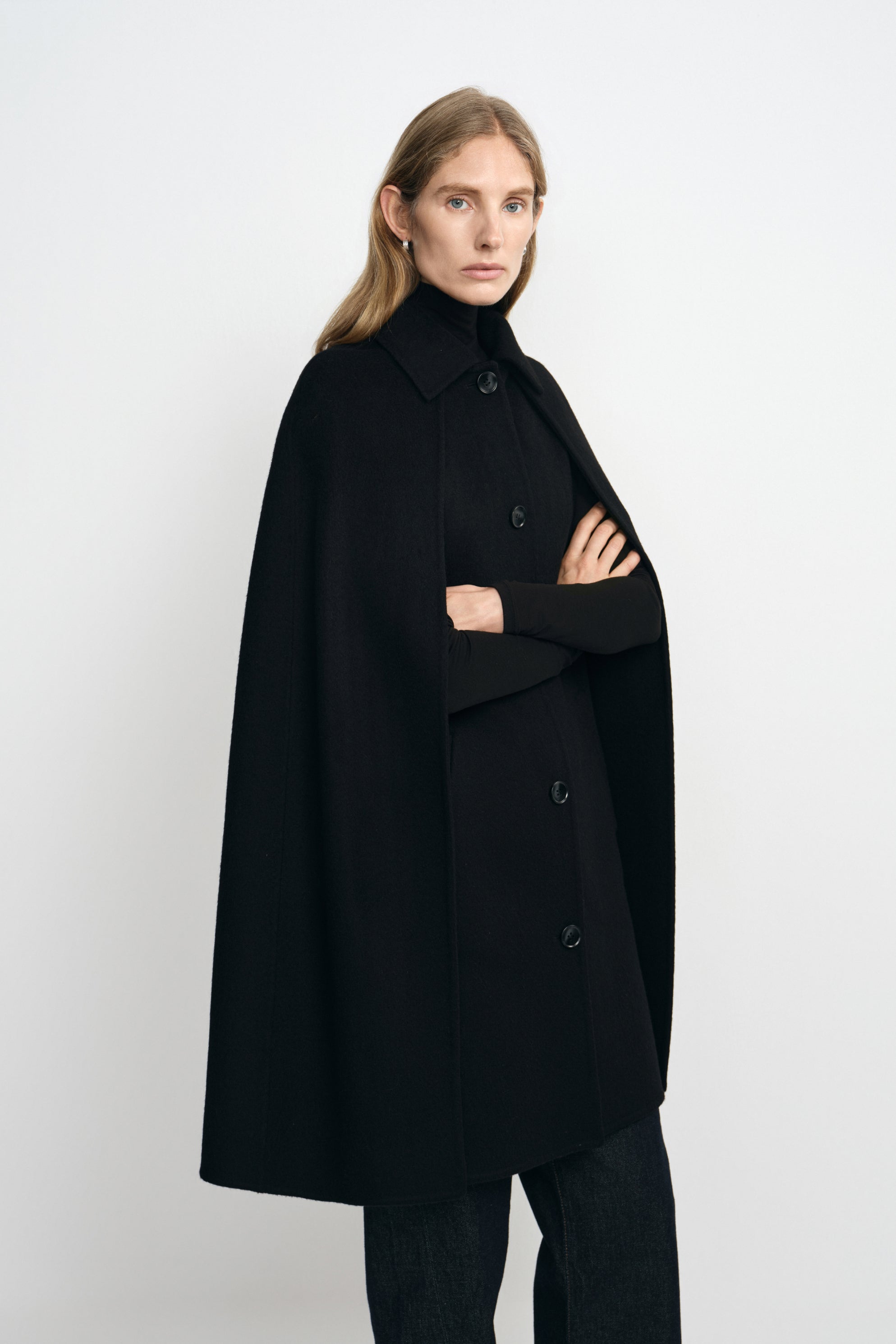 Three-quarter view of a model in a black cape coat, with folded arms emerging from the openings beneath the pointed collar and button front.