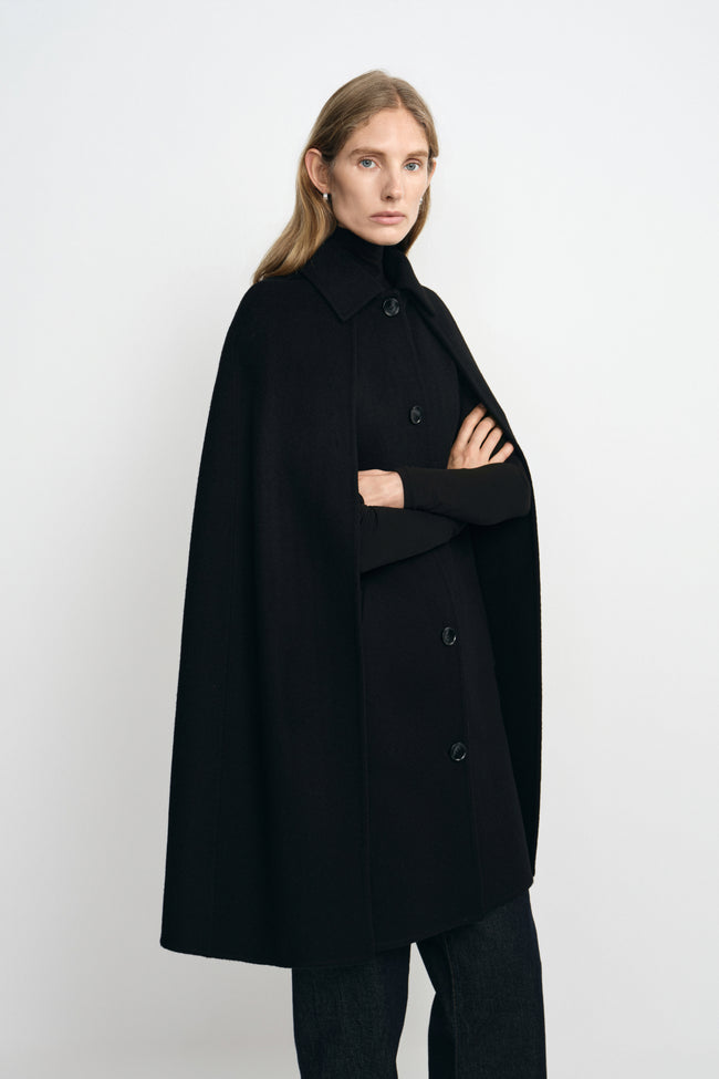 Three-quarter view of a model in a black cape coat, with folded arms emerging from the openings beneath the pointed collar and button front.