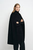 Three-quarter view of a model in a black cape coat, with folded arms emerging from the openings beneath the pointed collar and button front.