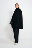 Side view of a model wearing a black cape coat with a wide hem, styled with dark trousers, pointed white shoes and a black handbag.