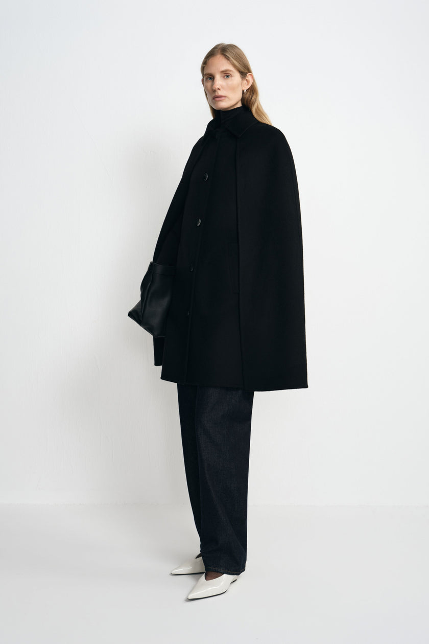 Side view of a model wearing a black cape coat with a wide hem, styled with dark trousers, pointed white shoes and a black handbag.