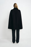 Back view of a model in a black cape coat, showing its broad A-line drape and center-back seam above dark trousers and white shoes.