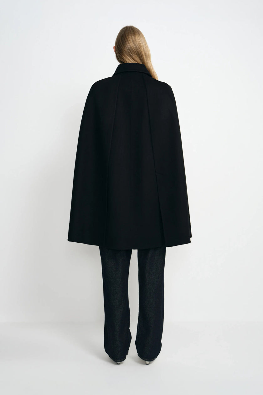 Back view of a model in a black cape coat, showing its broad A-line drape and center-back seam above dark trousers and white shoes.