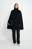 A model wearing a black wool-cashmere-blend cape coat with a pointed collar and button front, over a black turtleneck and dark trousers with white shoes.