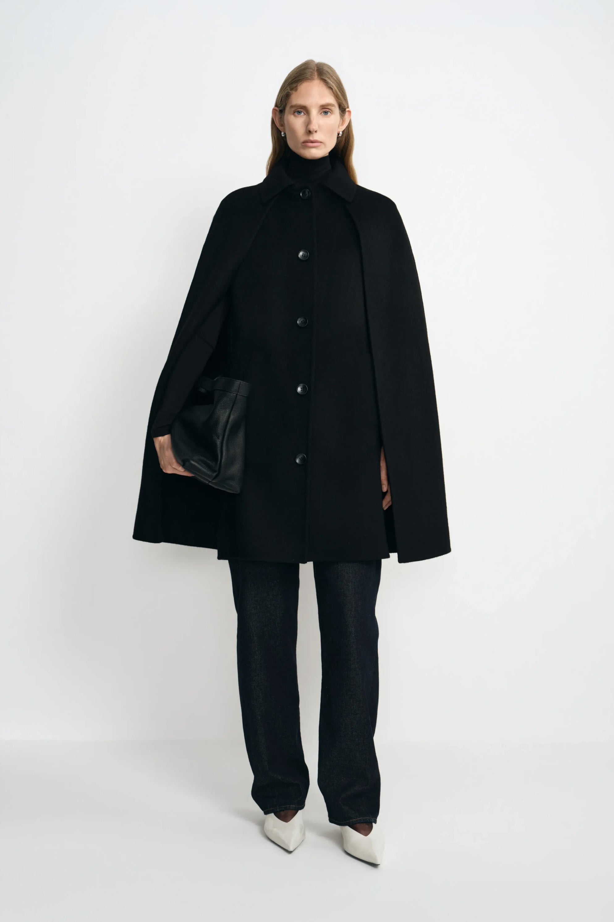 A model wearing a black wool-cashmere-blend cape coat with a pointed collar and button front, over a black turtleneck and dark trousers with white shoes.