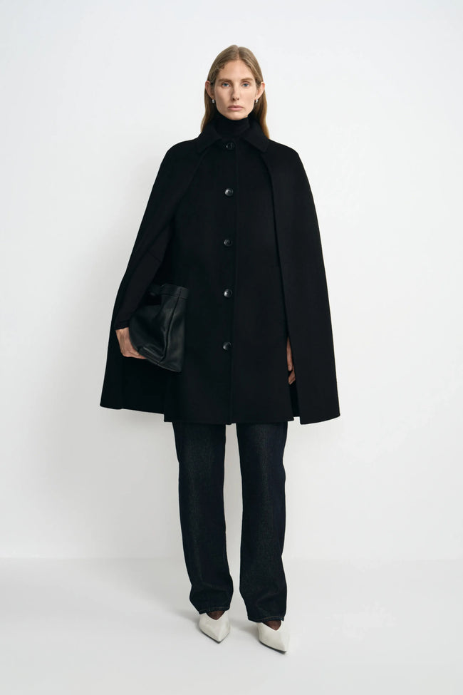 A model wearing a black wool-cashmere-blend cape coat with a pointed collar and button front, over a black turtleneck and dark trousers with white shoes.