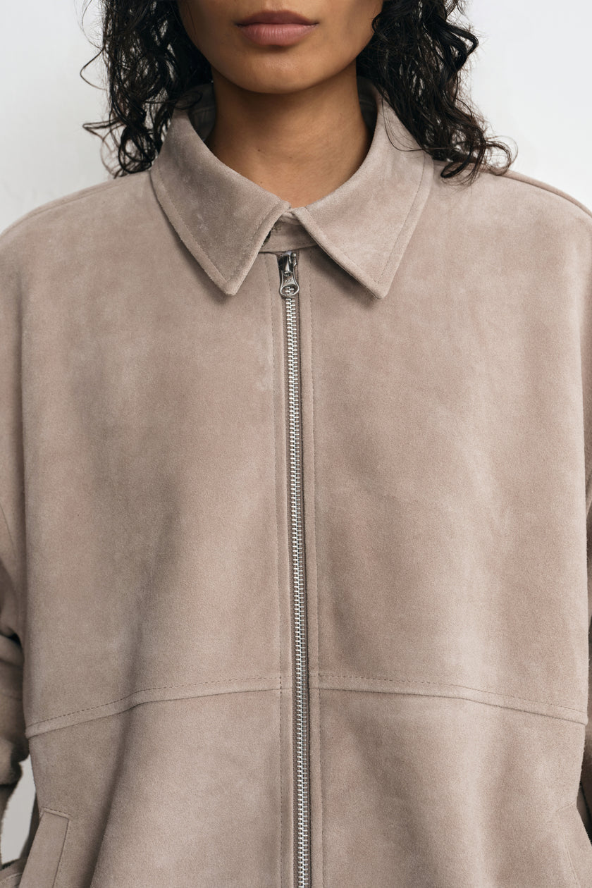 Close-up of a slate-grey jacket on a model, showing the napped surface, pointed collar, metal zipper and horizontal stitched panel seam.