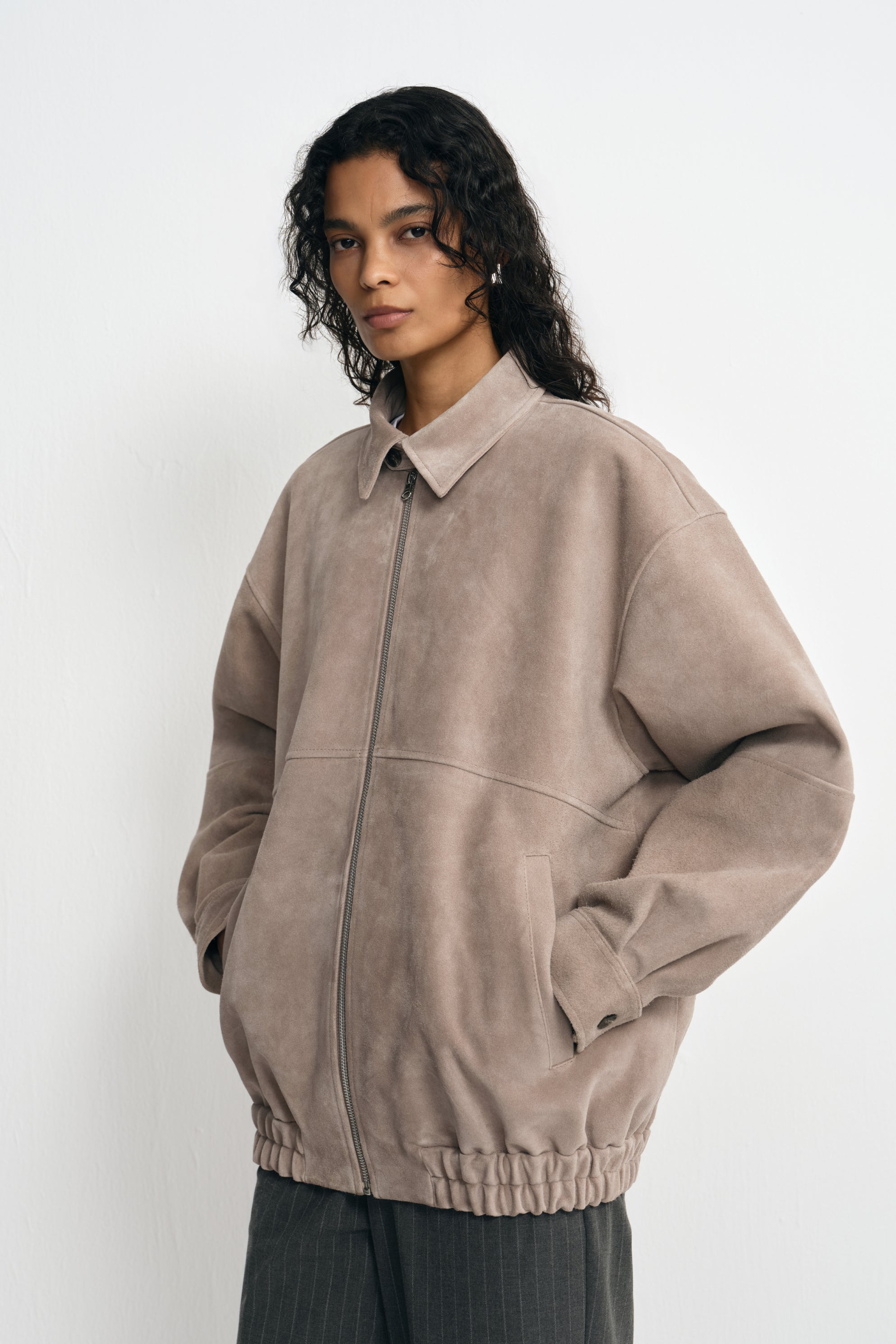 Three-quarter view of a model in a zipped slate-grey jacket, with hands in the pockets and full sleeves above a gathered hem and striped trousers.
