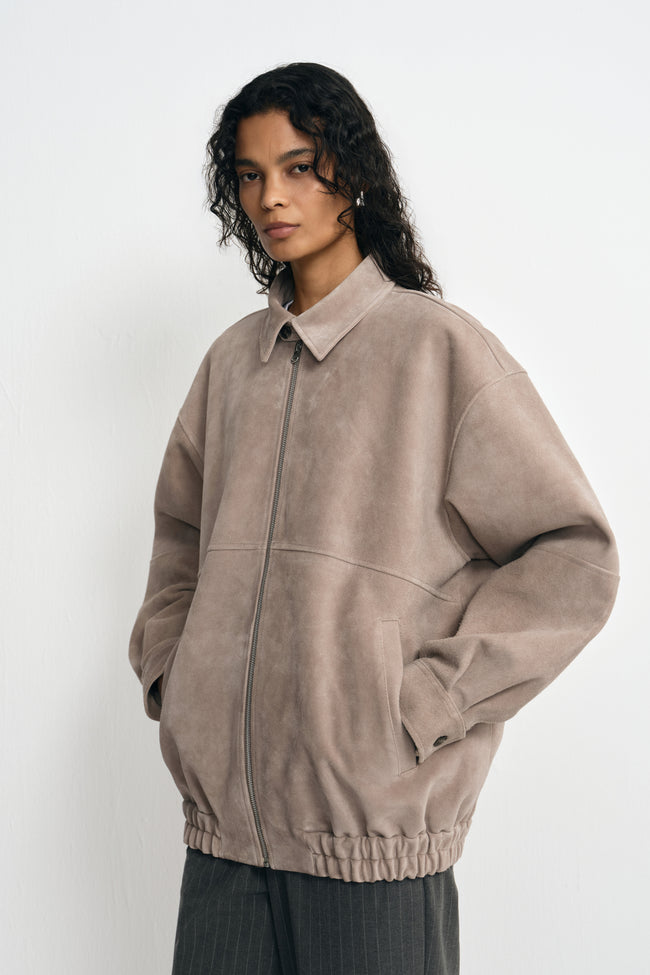 Three-quarter view of a model in a zipped slate-grey jacket, with hands in the pockets and full sleeves above a gathered hem and striped trousers.