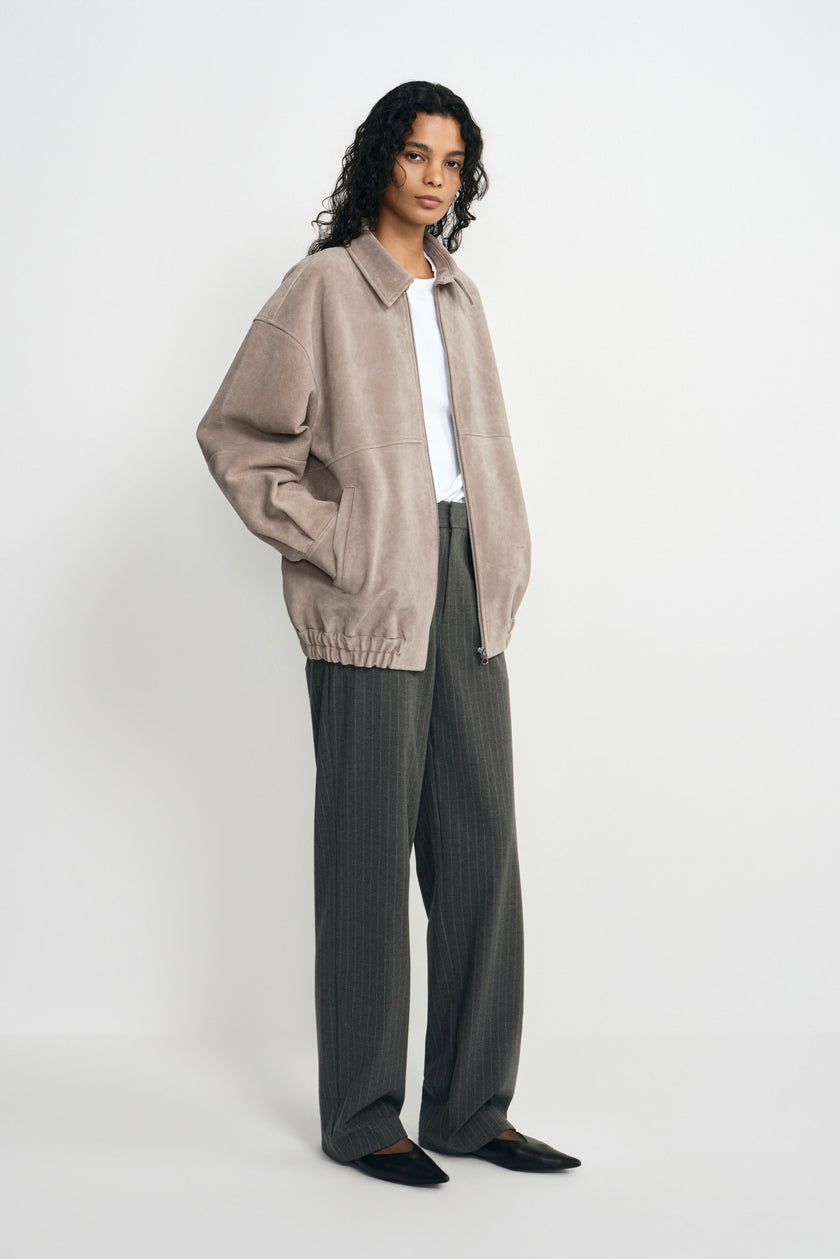 A model wearing a slate-grey calfskin jacket open over a white T-shirt and charcoal pinstriped trousers, with a gathered hem and black pointed shoes.