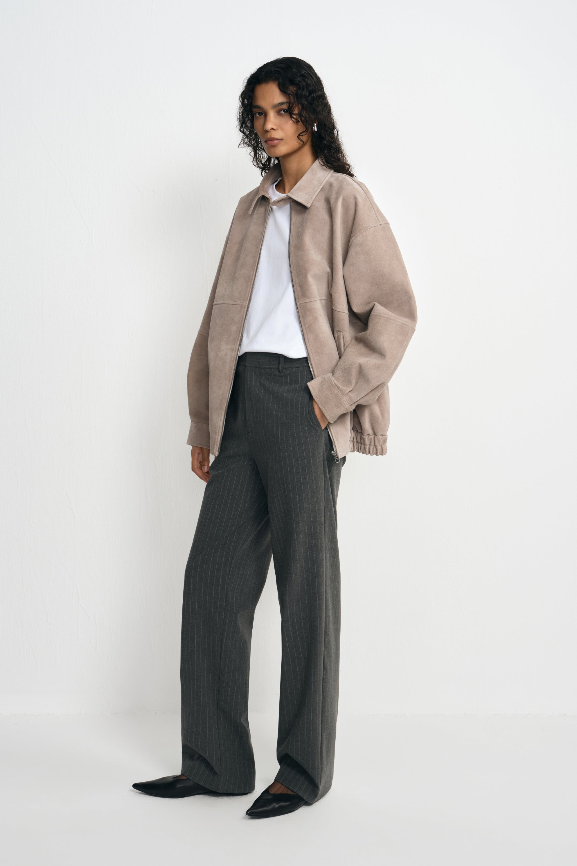 Three-quarter view of a model in an open slate-grey jacket with dropped shoulders, paired with a white T-shirt, charcoal striped trousers and black shoes.