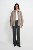 A model facing forward in a slate-grey jacket with a pointed collar and gathered hem, worn open over a white T-shirt with charcoal trousers and black shoes.