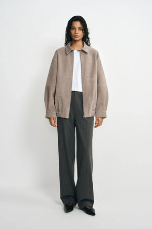 A model facing forward in a slate-grey jacket with a pointed collar and gathered hem, worn open over a white T-shirt with charcoal trousers and black shoes.