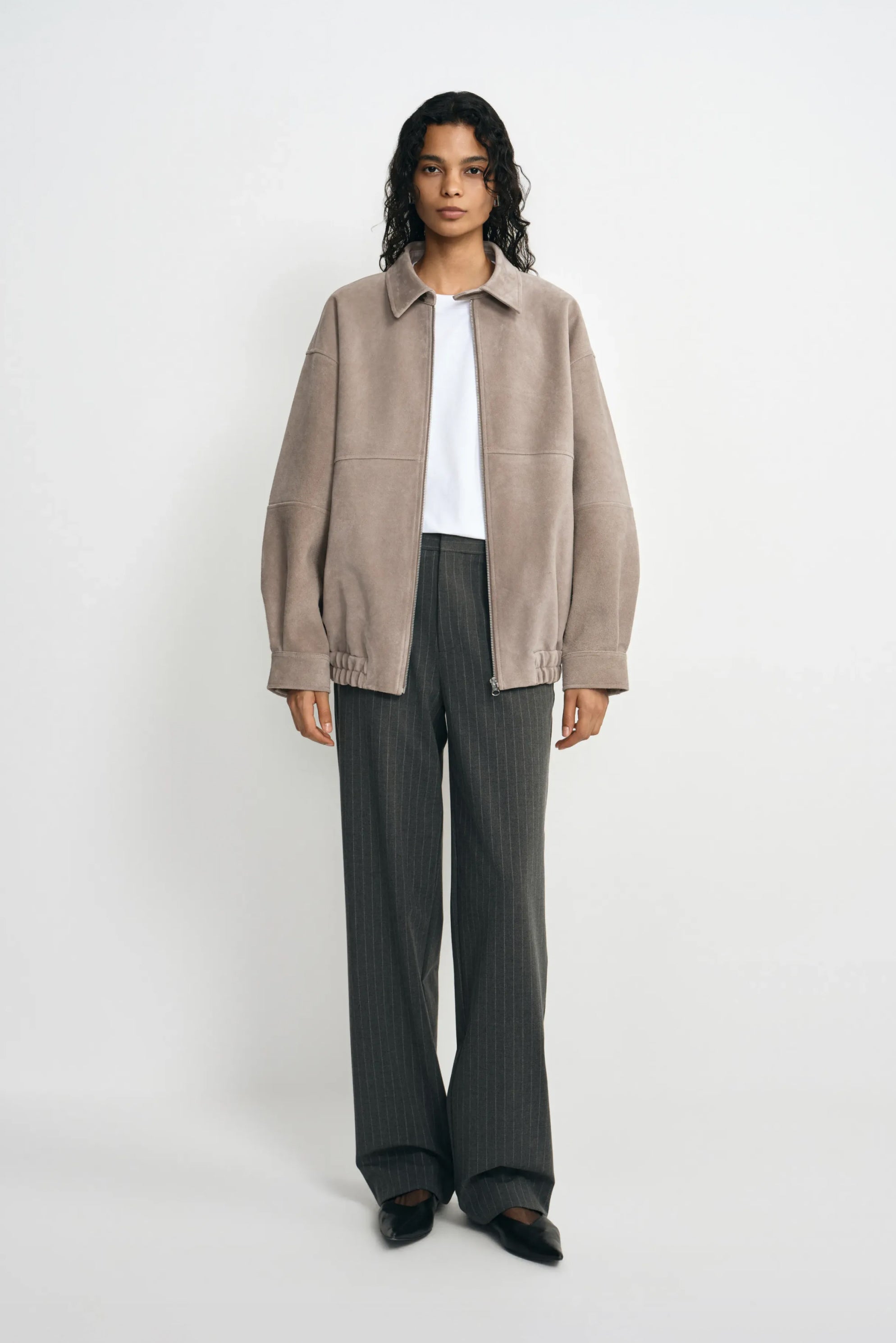 A model facing forward in a slate-grey jacket with a pointed collar and gathered hem, worn open over a white T-shirt with charcoal trousers and black shoes.