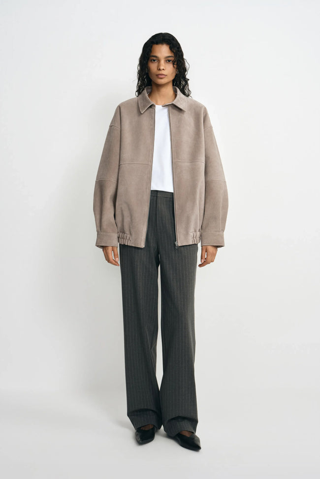 A model facing forward in a slate-grey jacket with a pointed collar and gathered hem, worn open over a white T-shirt with charcoal trousers and black shoes.