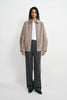A model facing forward in a slate-grey jacket with a pointed collar and gathered hem, worn open over a white T-shirt with charcoal trousers and black shoes.