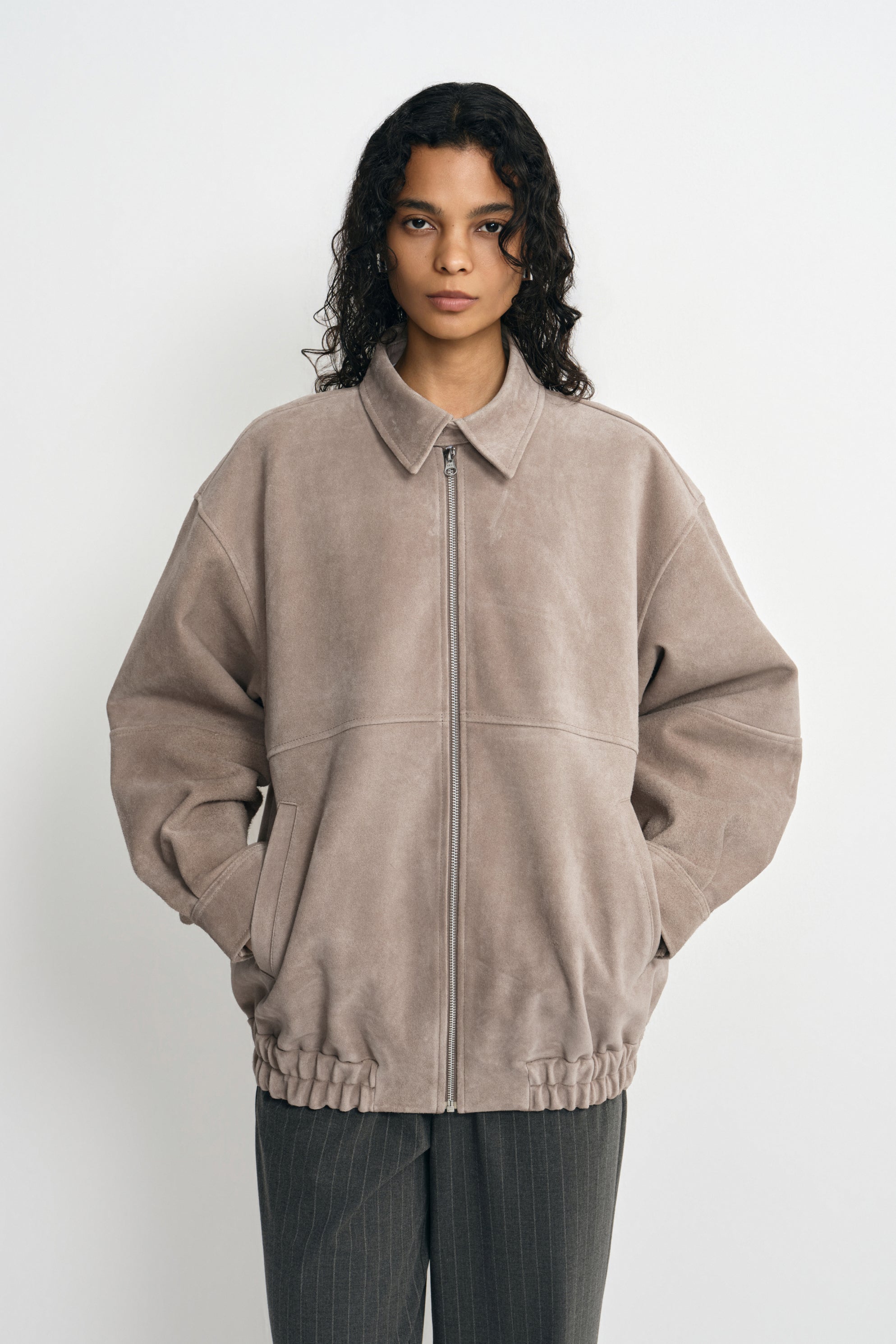 A model wearing a slate-grey calfskin jacket zipped to the pointed collar, with dropped shoulders and a gathered hem above charcoal pinstriped trousers.