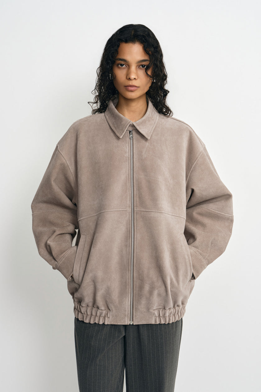 A model wearing a slate-grey calfskin jacket zipped to the pointed collar, with dropped shoulders and a gathered hem above charcoal pinstriped trousers.