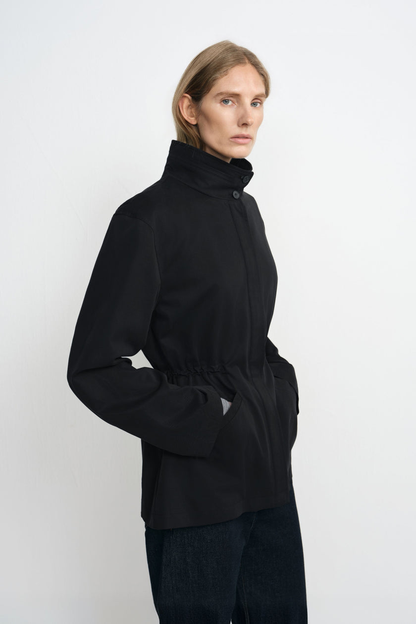 Three-quarter close-up of a model in a black jacket, showing the high buttoned collar and gathered drawstring waist with both hands in the pockets.