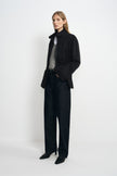 Angled view of a model wearing a black jacket with a raised collar and cinched waist, open over grey and white tops with dark jeans and black shoes.