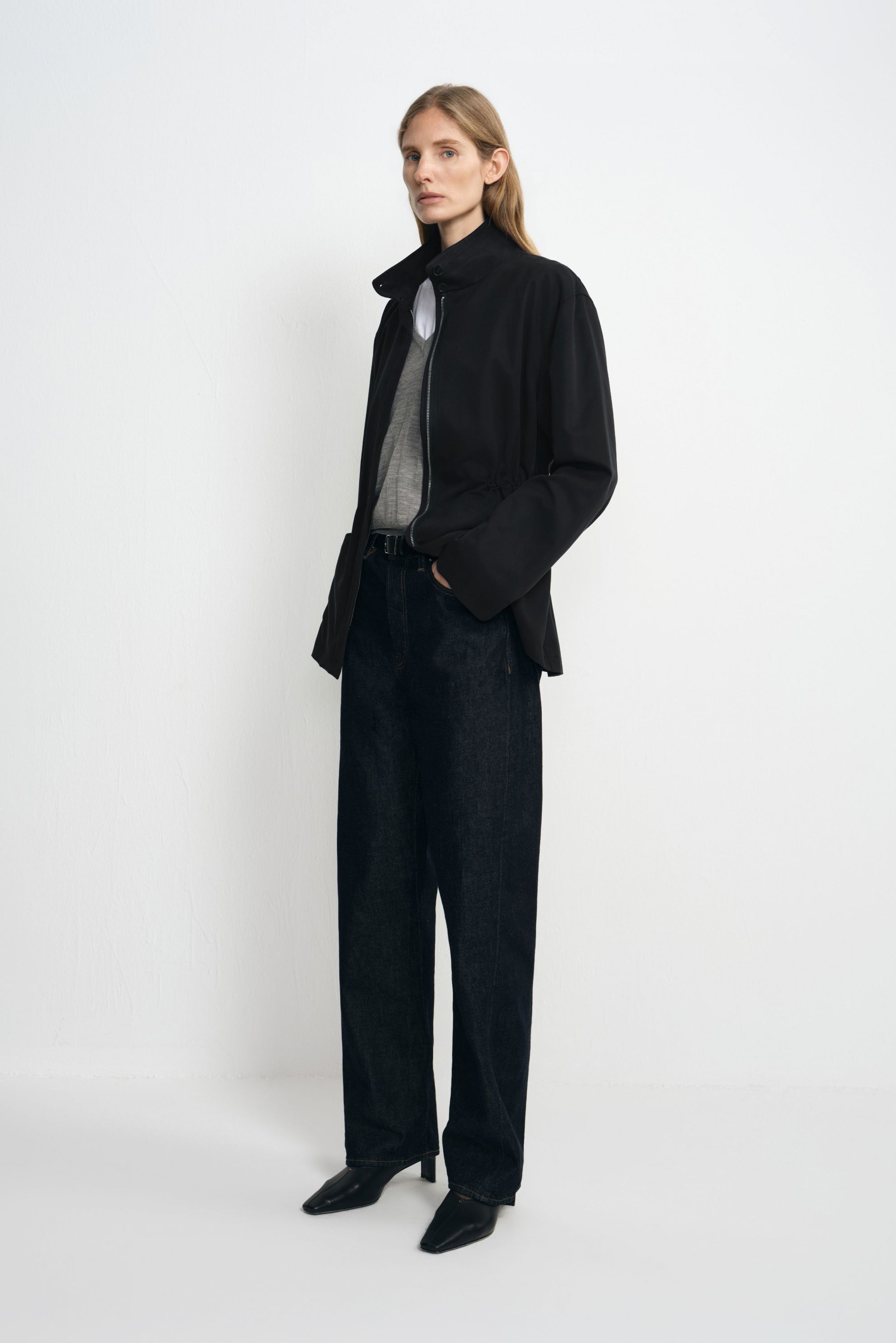 Angled view of a model wearing a black jacket with a raised collar and cinched waist, open over grey and white tops with dark jeans and black shoes.