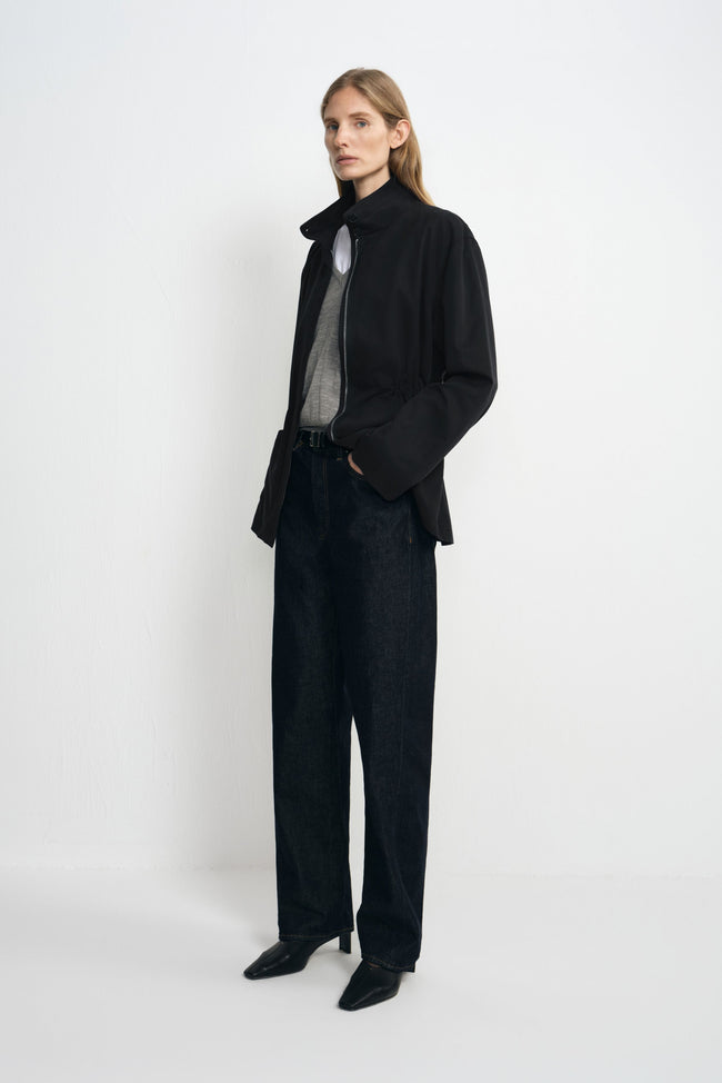 Angled view of a model wearing a black jacket with a raised collar and cinched waist, open over grey and white tops with dark jeans and black shoes.