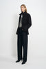 Angled view of a model wearing a black jacket with a raised collar and cinched waist, open over grey and white tops with dark jeans and black shoes.